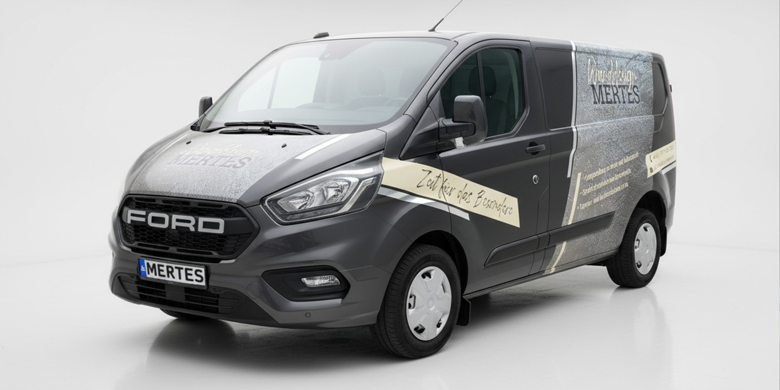 Ford Trnasit Transporter in silbergrau metallic mit Werbebeschriftung in Creme-Grau seitlich zu sehen. Grafische Verzierung in Chrom als Highlight.