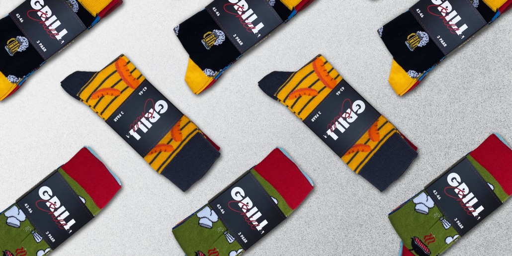 Produktfotografie versetzt angeordneten Verpackungen mit Grillmotiven auf den Socken. Schwarze Banderole mit GRILL & Chill Aufschrift.