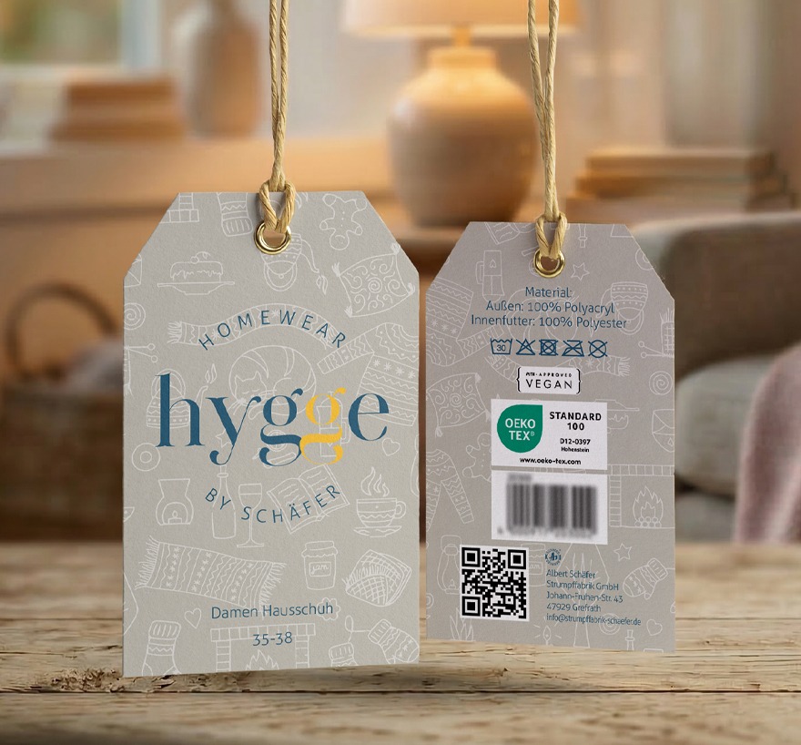 Hangtag haengend fotografiert mit dezenten weißen Icons Symbolen Motiven die gemuetlichkeit udn eine hyggelige Atmosphaere Ausstrahlen, Schal, Teppich, Kerze, Buch, Katze, Pullover, Tee Handschuhe. Das Hangtag ist von beiden Seiten dargestellt, das Logo ist blau und gelb.