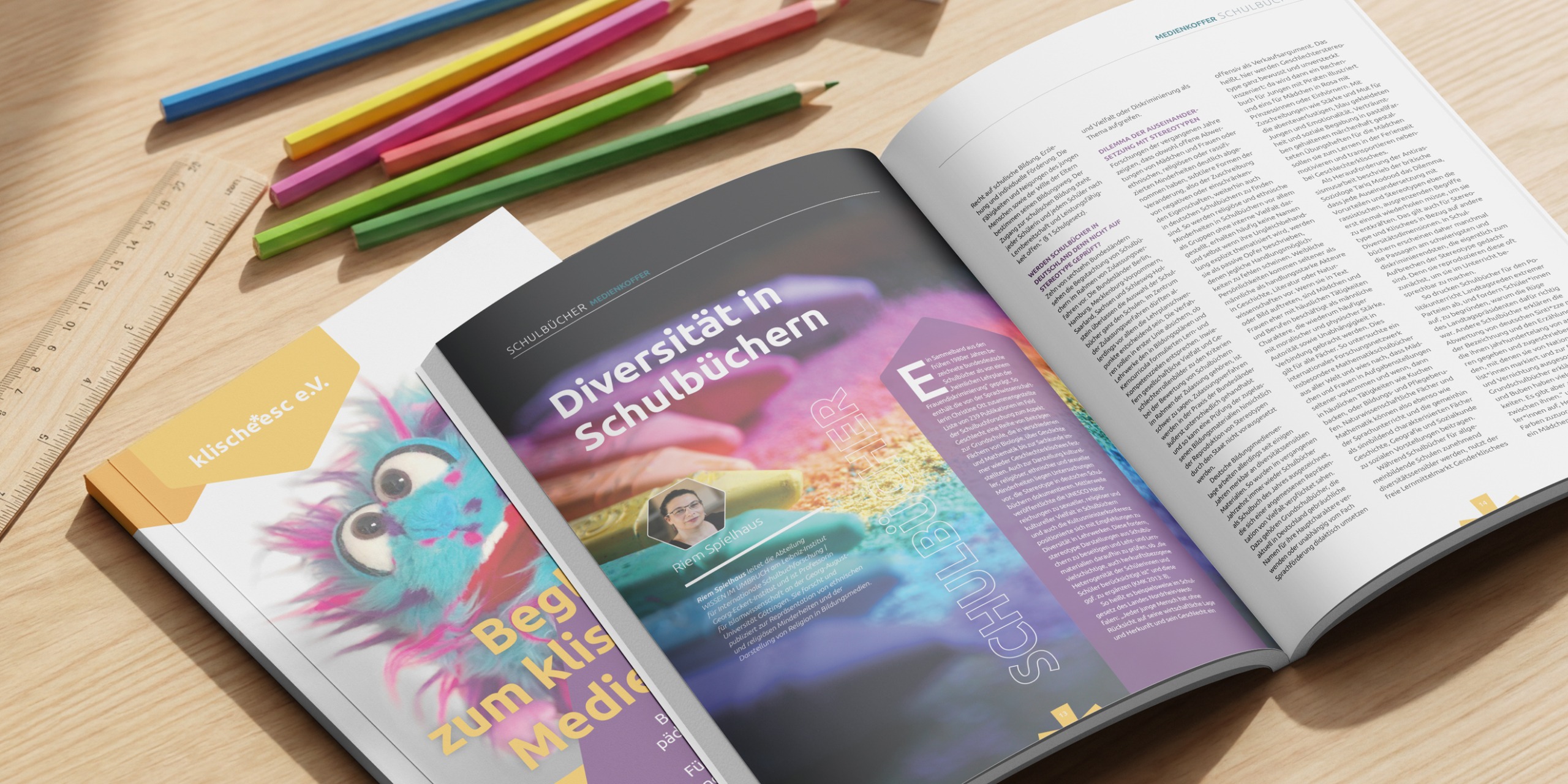 Aufgeklappte Broschueren des Medienkoffer Begleithefts zum klische*esc Medienkoffer. Die Titelseite des Begleithefte ist unter dem aufgeschlagenen Inneteil des Heftes zu sehen. Sichtbar ist der Artikel Diversitaet in Schulbuechern. Liegend auf einem hellen Holztisch, neben der Broschuere liegen Buntstifte und ein Lineal.