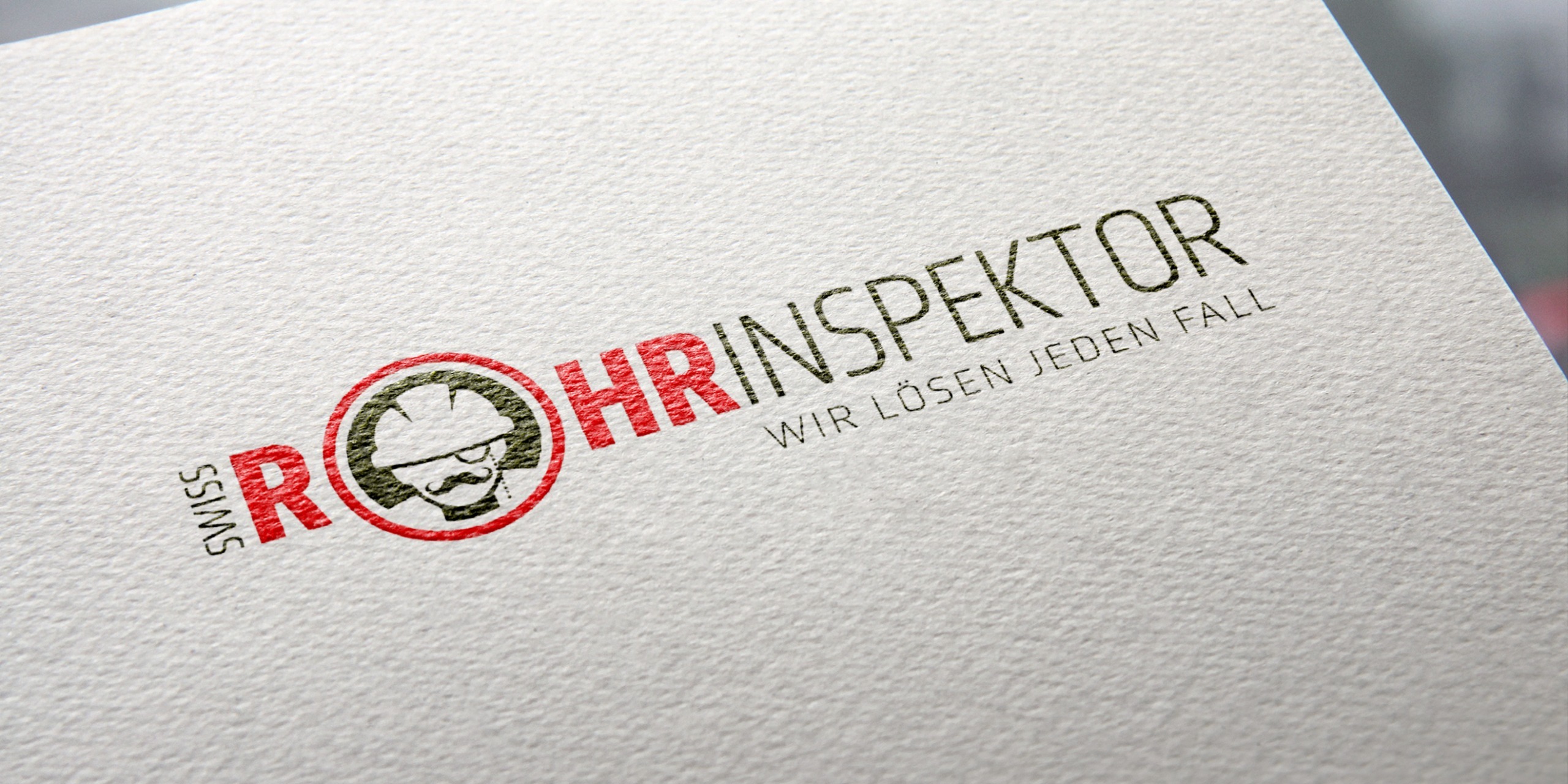 Das Logo der Firma swiss Rohrinspektor mit Claim darunter „Wir Loesen jeden Fall“ dargestellt auf einem ehllen strukturiertem Papieruntergrund. Leicht schräge perspektive.