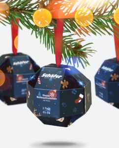 Produktfotografie einer weihnachtlichgemusterten Verpackung in Form einer Kugel mit weihnachtlichem Socken Inhalt, hängend an einem Tannenast.